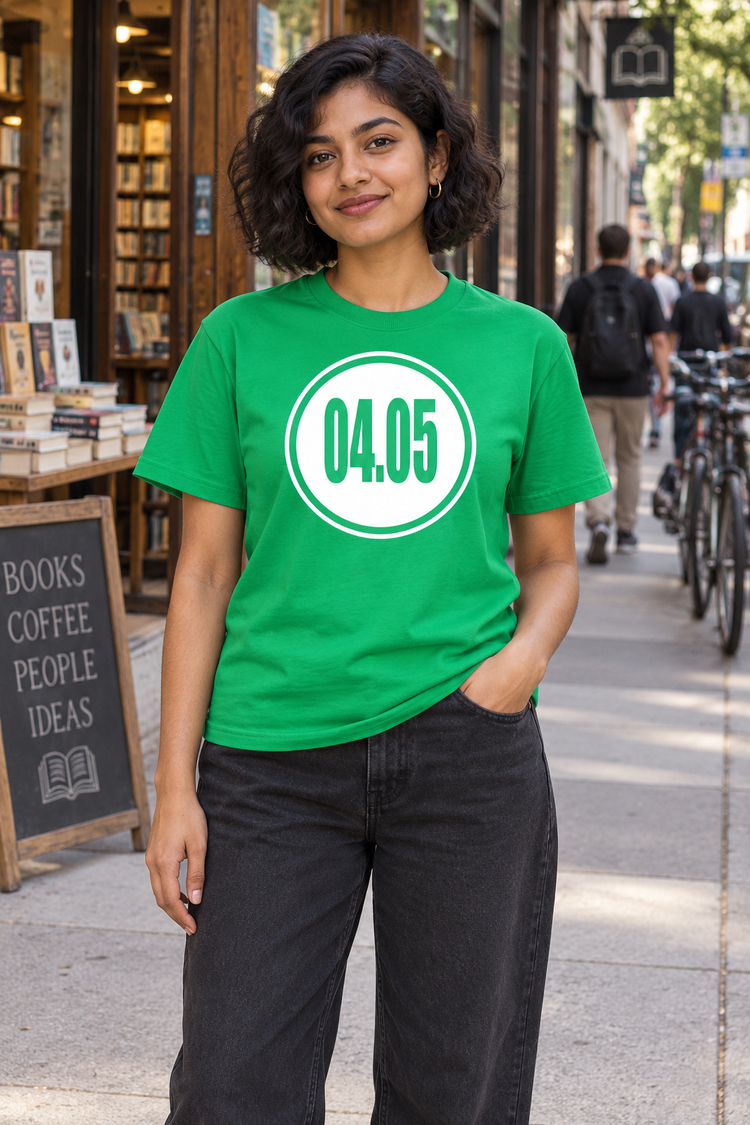 AI-generated South Asian woman age 24, medium brown skin, petite build, wavy dark bob wearing Women’s 04.05 Bordered Circle Classic Tee — Irish Green; busy downtown bookstore entrance with pedestrians and bike racks