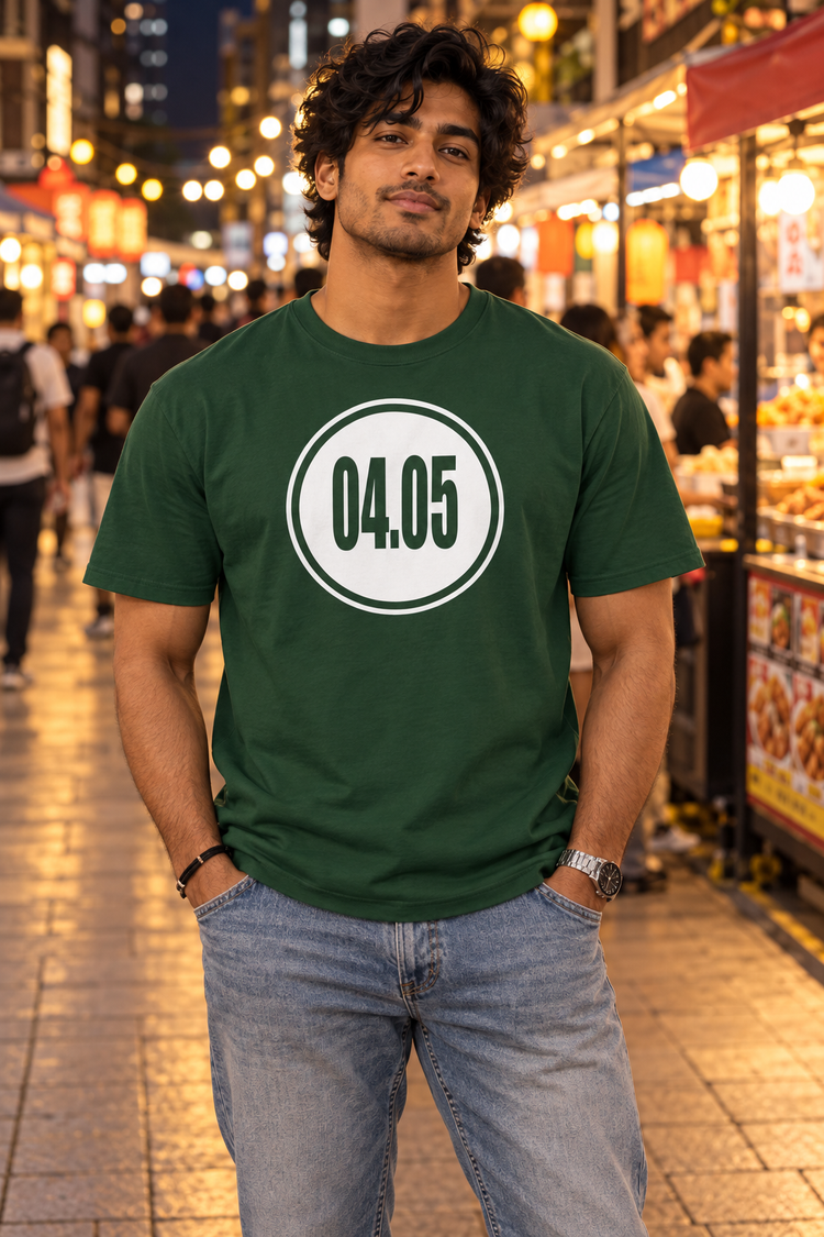 AI-generated South Asian man age 24, brown skin, broad athletic build, medium-length wavy hair wearing Men’s 04.05 Bordered Circle Classic Tee — Forest Green; busy downtown evening food market, warmly lit stalls and walking crowds