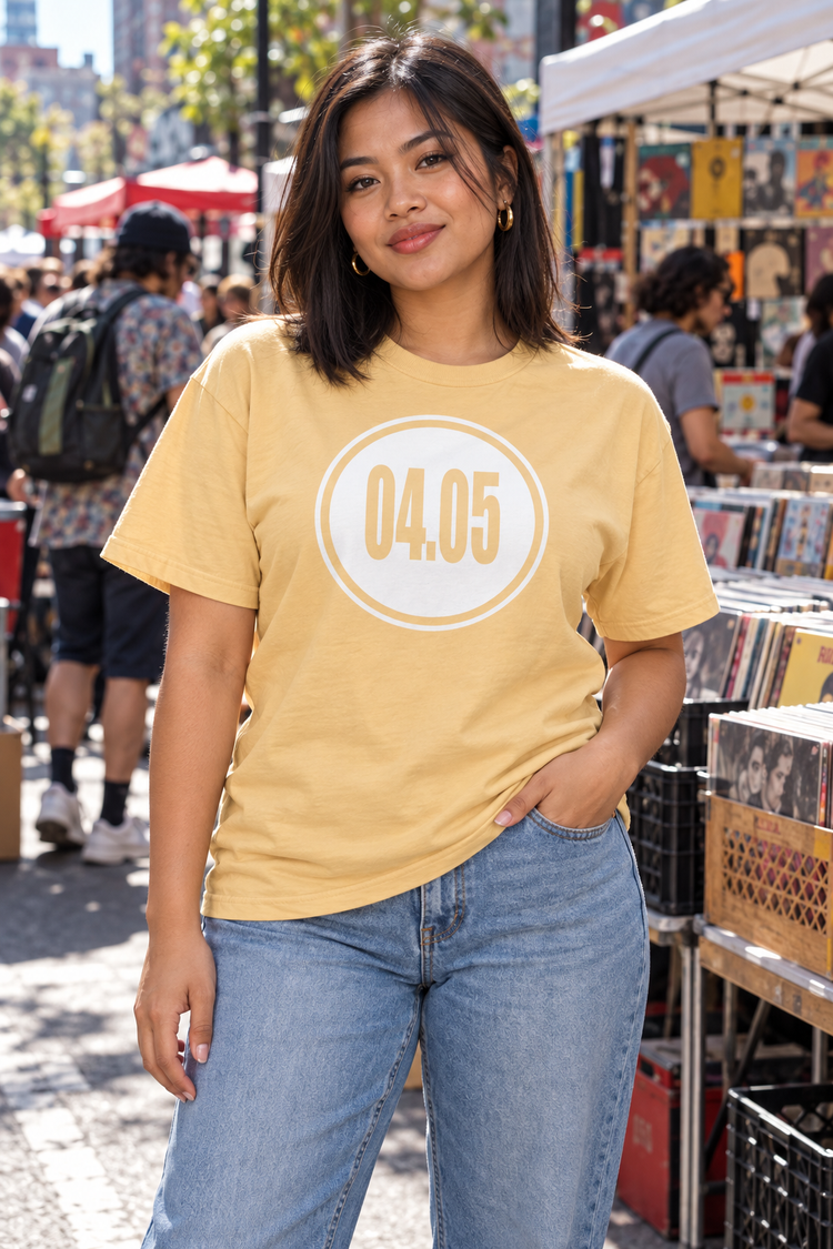 AI-generated Southeast Asian woman age 25, warm tan skin, curvy build, straight shoulder-length hair wearing Women’s 04.05 Bordered Circle Classic Tee — Yellow Haze; busy downtown outdoor record market with shoppers and colorful stalls