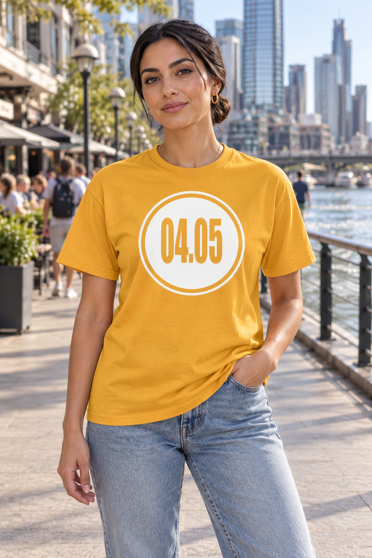 AI-generated Middle Eastern woman age 28, olive skin, athletic build, dark hair tied in low bun wearing Women’s 04.05 Bordered Circle Classic Tee — Gold; busy downtown riverside boardwalk with pedestrians, cafes and skyline
