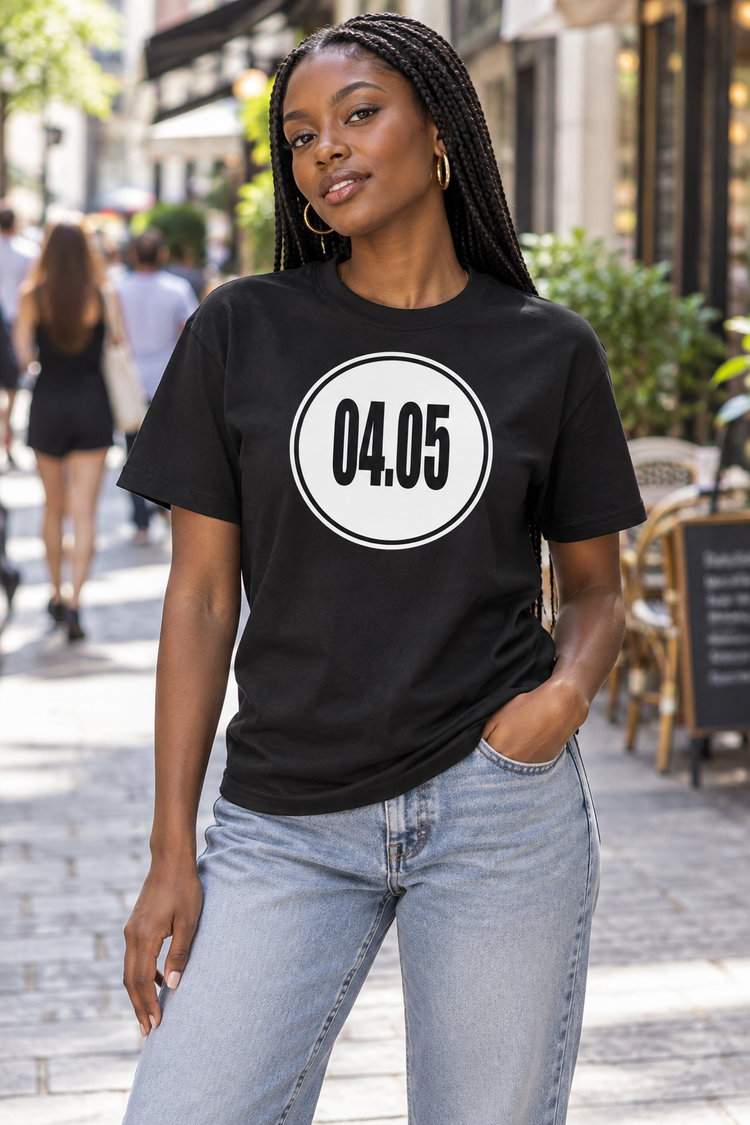 AI-generated Black woman age 23, deep brown skin, slim athletic build, long braids behind shoulders wearing Women’s 04.05 Bordered Circle Classic Tee — Black; busy downtown pedestrian shopping street with cafe terraces